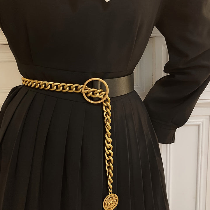 A close-up of a black pleated dress with a wide black belt featuring a prominent gold chain link and a hanging gold medallion, worn by a model.