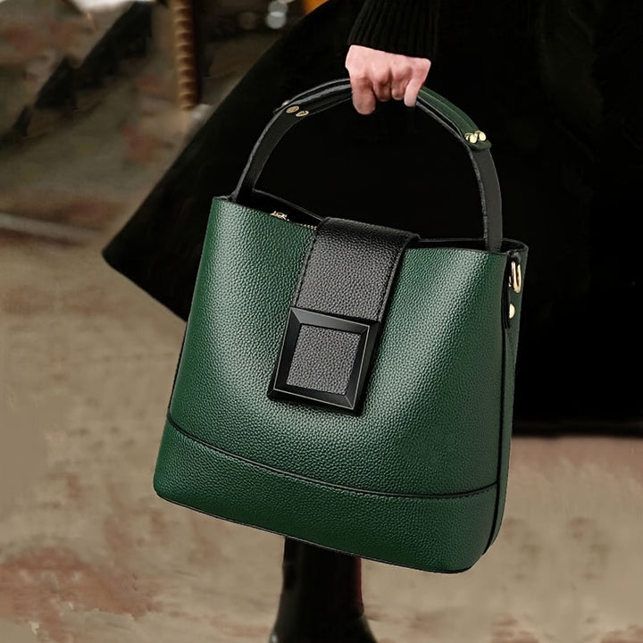 A dark green pebbled leather bucket handbag with black trim and a black front flap accent, held by a person wearing a black dress in an indoor setting.